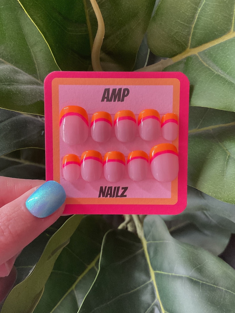 Pu&ograve; includere: Un set di dieci unghie finte rosa con un design bicolore arancione e rosa. Le unghie sono esposte su una carta rosa e arancione con il testo "AMP NAILZ".