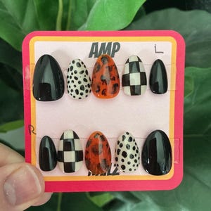 May include: A set of ten press-on nails in various designs. The nails feature glossy black, dalmatian print, orange tortoise shell, and black and white checkered patterns. The card reads "AMP" in white text.