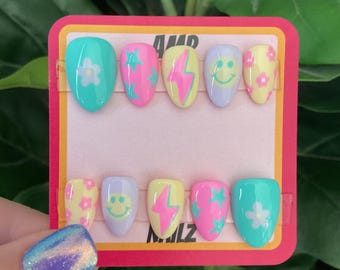 May include: A set of 8 colorful fake nails with different designs. The designs include flowers, stars, lightning bolts, and smiley faces. The nails are on a pink and red card with the text "AMP NAILS".