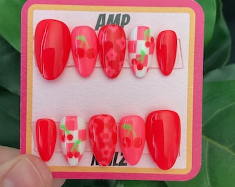 May include: A set of ten red press-on nails with different cherry and floral designs. The nails are arranged on a pink and white card with the text "AMP" and "MILZ".