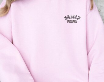 Könnte beinhalten: Hellrosa Sweatshirt mit dem Text "DOODLE MAMA" in dunkelvioletter, bogenförmiger Schrift. Das Sweatshirt hat lange Ärmel und einen Rundhalsausschnitt. Der Stoff wirkt weich und bequem.