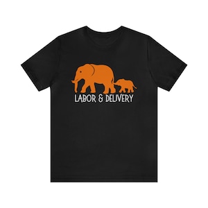 May include: Black t-shirt featuring an orange graphic of an adult elephant and a baby elephant walking together. The text "LABOR & DELIVERY" is printed in white below the elephants. The shirt is made of a soft material.