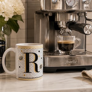 May include: A white ceramic mug featuring a large black letter R centred on a gold and black honeycomb pattern with small floral accents. The mug has a gold-trimmed rim and base, and is positioned next to a stainless steel espresso machine.