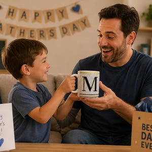 May include: A white ceramic mug featuring a black letter M centered on a gold and black honeycomb pattern. The mug has a gold rim and base, with a handle on the side. The design is clean and functional, showcasing a bold typographic element.