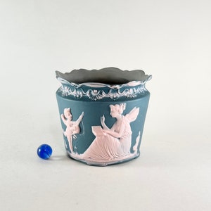 May include: A blue and pink ceramic planter with a scalloped rim and a raised design of two figures. The figures are in a pastel pink color and are depicted in a classical style.