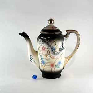 May include: A white ceramic teapot with a black and gold dragon design. The teapot has a gold trim and a black handle.