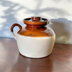 May include: A white and brown ceramic crock with a lid. The crock has a brown glaze on the top half and a white glaze on the bottom half. The crock has a handle on the side.