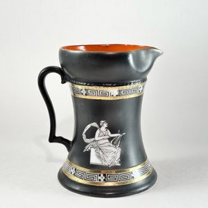 May include: Black ceramic pitcher with a gold and white Greek key design and a white silhouette of a woman playing a lyre. The pitcher has an orange rim.