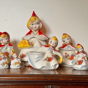 Pode incluir: Um conjunto de chá de cerâmica vintage com um design de menina com capuz vermelho. O conjunto inclui uma chaleira, um jarro de creme, um açucareiro e dois saleiros e pimenteiros. As peças são decoradas com padrões florais e um esquema de cores vermelho, branco e amarelo.