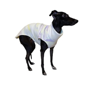 May include: A black Italian Greyhound dog wearing a sleeveless turtleneck sweater. The sweater is white with faint, soft-toned pastel tie-dye patterns in shades of light yellow and purple. The dog is standing in profile, facing toward the right side.