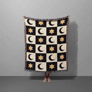 May include: A woven throw blanket featuring a repeating checkerboard pattern of black and cream squares. The black squares contain a cream crescent moon, while the cream squares feature a tan six-pointed star. The blanket has fringed edges.