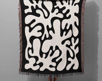 May include: Black and white woven throw blanket with a large abstract pattern of organic shapes. The blanket has a fringe border.