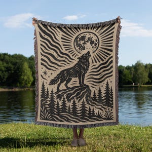 Peut inclure: Un plaid tissé présentant un motif graphique noir et beige représentant un loup hurlant à la pleine lune. L'illustration comprend des lignes ondulées stylisées, une forêt de pins à la base et des bords à franges en haut et en bas.