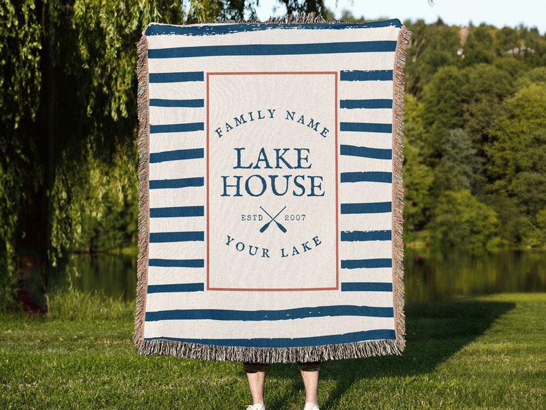 以下が含まれることがあります： 青と白のストライプの織り毛布。毛布には、「FAMILY NAME LAKE HOUSE ESTD 2007 YOUR LAKE」というテキストが書かれた茶色の長方形の中に、パーソナライズされたデザインが施されています。