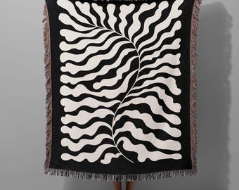 May include: A rectangular woven throw blanket featuring a high-contrast black and off-white abstract botanical leaf pattern. The design displays a central stem with wavy, organic fronds extending outwards. The edges are finished with a fringed border.