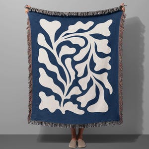 Op de afbeelding: Een blauwe geweven deken met een wit abstract bloemenontwerp. De deken heeft aan alle kanten franjes.