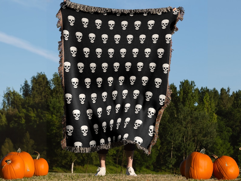 May include: A black throw blanket with a white skull pattern. The blanket is being held up in front of a person standing in a field with pumpkins.