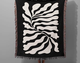 May include: Black and white woven throw blanket with a large abstract leaf design. The blanket has a fringed edge.