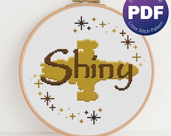 May include: A cross stitch pattern featuring the word "Shiny" in a gold and brown color scheme. The word is surrounded by small gold stars.