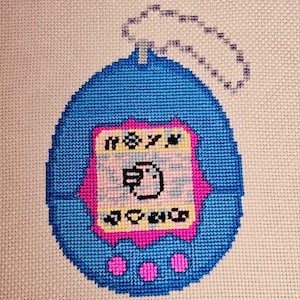 May include: A blue and pink cross-stitch of a Tamagotchi, a popular virtual pet toy from the 1990s. The Tamagotchi has a screen with a small bird on it and three buttons on the bottom.