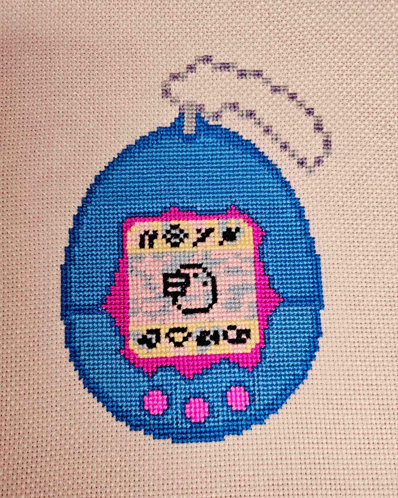 May include: A blue and pink cross-stitch of a Tamagotchi, a popular virtual pet toy from the 1990s. The Tamagotchi has a screen with a small bird on it and three buttons on the bottom.