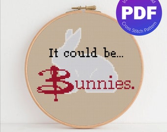 May include: A cross stitch pattern featuring a white bunny with the text "It could be... Bunnies." The bunny is lying down with its head turned to the side. The text is in black and the bunny is outlined in black. The background is a light beige color.