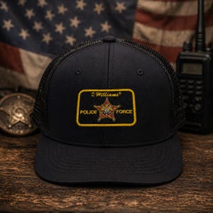 May include: Black trucker hat with a mesh back and a patch that reads "Williams Police Force." The patch features a star emblem with the words "Police" and "Force" on either side. An American flag is in the background.