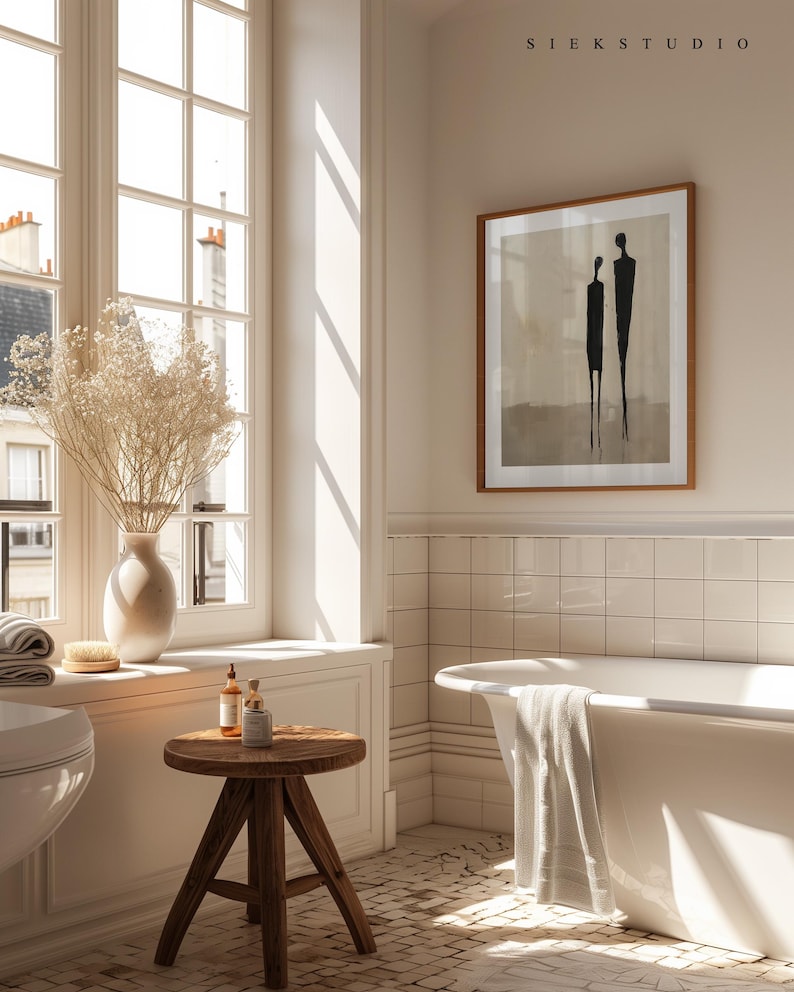 May include: A white bathtub with a towel draped over the edge, a wooden stool with two bottles and a jar on top, and a framed print of two figures on a beige background. The print is titled "SIEKS STUDIO".