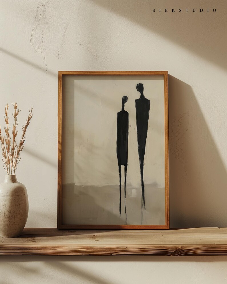 May include: A framed abstract painting of two black figures standing side-by-side on a light beige background. The painting is in a wooden frame and is hanging on a wooden shelf.