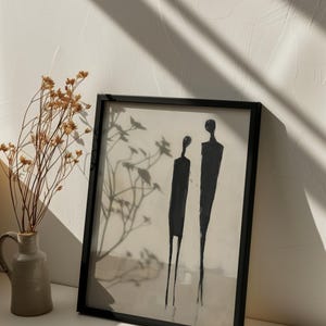 May include: A black framed print with a minimalist abstract painting of two figures in black against a white background. The print is hanging on a white wall with a vase of dried flowers in the foreground.