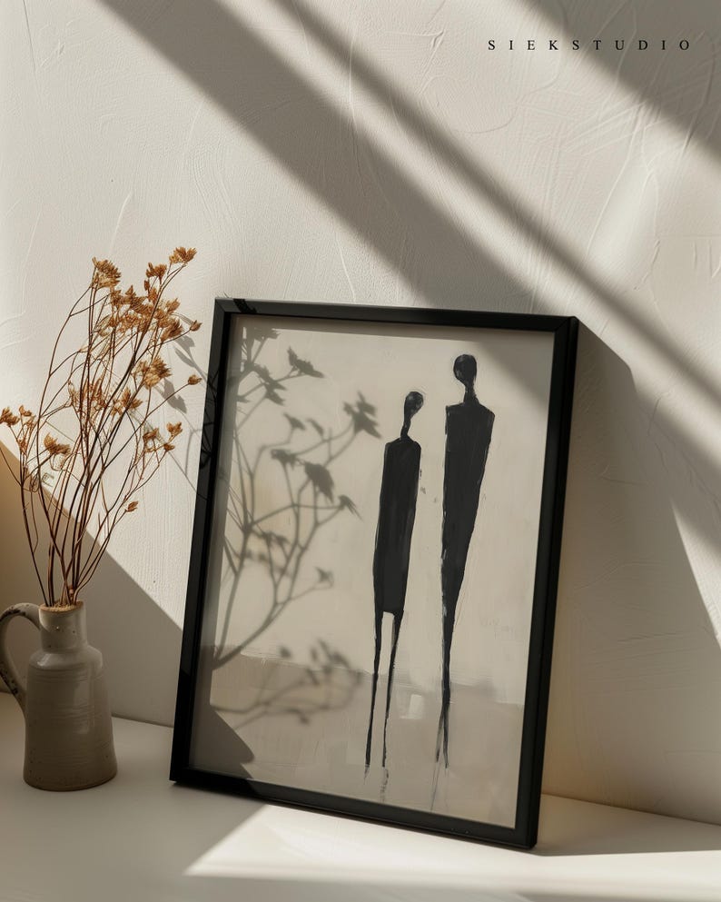 May include: A black framed print with a minimalist abstract painting of two figures in black against a white background. The print is hanging on a white wall with a vase of dried flowers in the foreground.