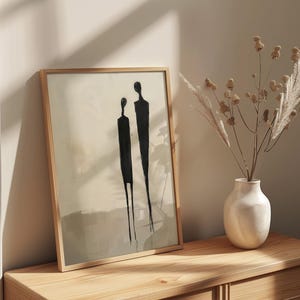 May include: A framed abstract art print with two black figures on a beige background. The print is hanging on a light wood dresser with a white vase and dried flowers.