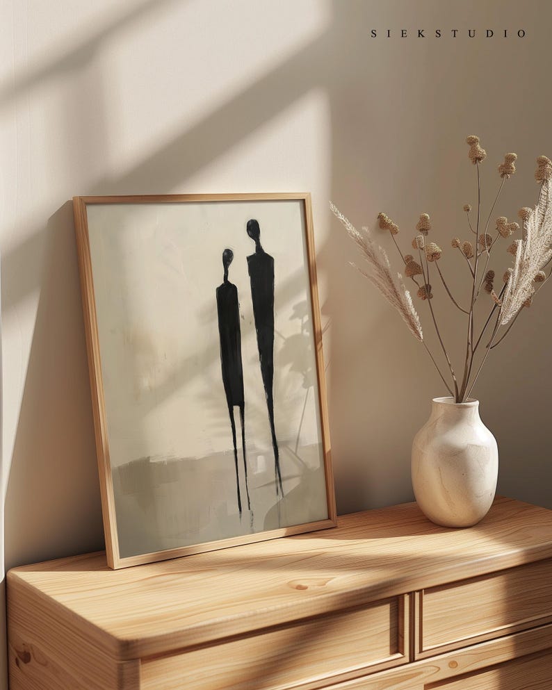 May include: A framed abstract art print with two black figures on a beige background. The print is hanging on a light wood dresser with a white vase and dried flowers.