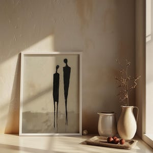 May include: A white framed print of a minimalist black and white abstract art piece depicting two figures standing side by side. The print is on a white shelf with a white ceramic pitcher, a beige ceramic pitcher, and a wooden tray with three brown spheres.