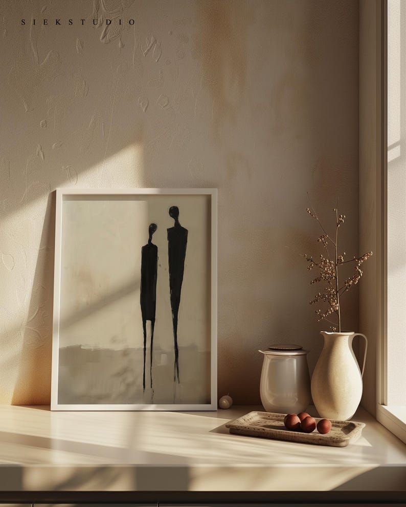 May include: A white framed print of a minimalist black and white abstract art piece depicting two figures standing side by side. The print is on a white shelf with a white ceramic pitcher, a beige ceramic pitcher, and a wooden tray with three brown spheres.