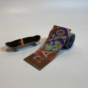 Puede incluir: Un monopatín de dedo en miniatura con una parte superior de cinta de agarre negra y una rampa curva con textura de metal oxidado, letras estilo grafiti moradas y verdes, y el texto blanco RA parcialmente visible en la sección inferior.