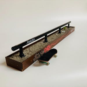 Puede incluir: Un obstáculo de skatepark en miniatura para fingerboard que presenta una larga barandilla de metal negro montada sobre una base rectangular de madera con arte de grafiti en el lateral. Un pequeño finger skate negro con ruedas verdes descansa contra la base.