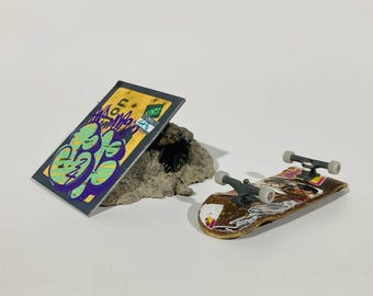 May include: A fingerboard skateboard with colorful stickers and a gray truck. The skateboard is on a white surface next to a small, gray rock and a yellow and green piece of cardboard with graffiti.