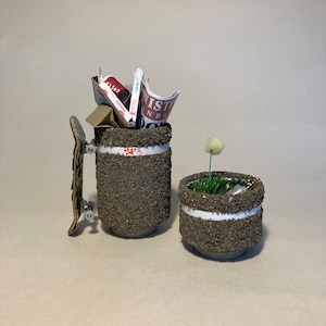 May include: Two small, textured, grey planters. One planter is filled with green grass and a white flower. The other planter has a skateboard and a stack of paper inside.