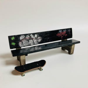 May include: A miniature black park bench with a graffiti-covered backrest featuring white bubble lettering, red script text, and two green circular stickers. A small black finger skateboard with light-colored wheels is positioned in front of the bench.