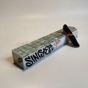 May include: A rectangular fingerboard obstacle featuring a white concrete-style block with a tiled top surface. The side displays black graffiti text reading SINISTER and red markings. A small black finger skateboard rests on the edge.