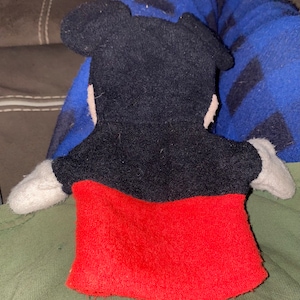 May include: A black and red plush hand puppet of Mickey Mouse. The puppet has a black face with large ears and a red shirt.