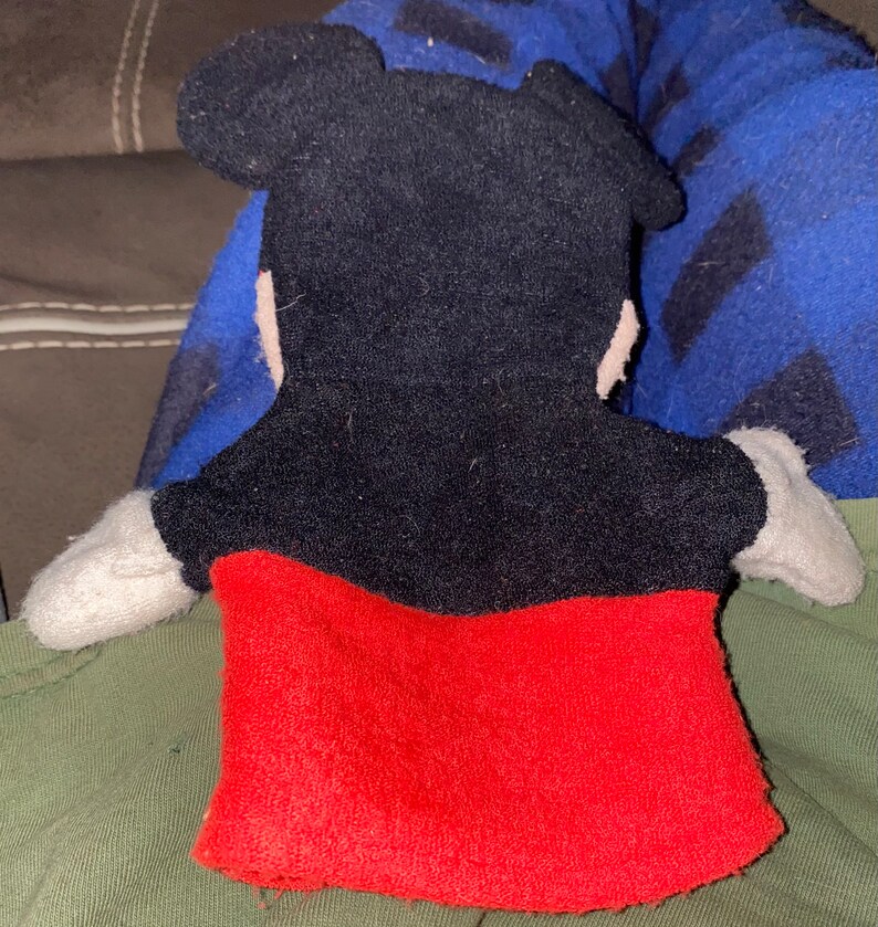 May include: A black and red plush hand puppet of Mickey Mouse. The puppet has a black face with large ears and a red shirt.