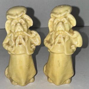 May include: A pair of yellow ceramic salt and pepper shakers shaped like cartoon dogs wearing hats. The dogs are holding a musical instrument in their right hand.