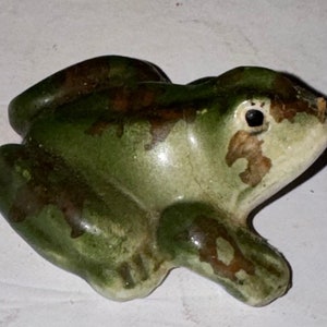 May include: A ceramic frog figurine with a green and brown mottled glaze. The frog has a detailed face with a black eye and a slightly open mouth.