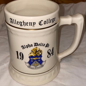 May include: White ceramic beer stein with a gold trim and the words "Allegheny College" and "Alpha Delta Pi 1984" printed on it. The stein features a blue and gold crest with a lion and the words "Fraterniy Amlnois".