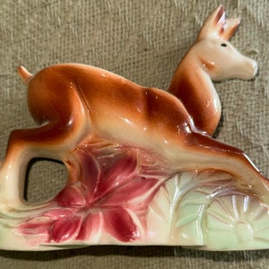 May include: A ceramic figurine of a brown deer leaping over a pink flower and green leaves.