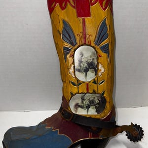 May include: A decorative cowboy boot figurine. The boot features a yellow shaft with red and blue accents, and a black heel. It has a spur and two oval images of cowboys on horseback.