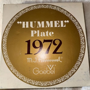 May include: A vintage Hummel plate box from 1972. The box features a gold-colored circular design with the words "HUMMEL Plate 1972" in a serif font. The box also includes the Goebel logo.