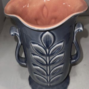 May include: A ceramic vase with a gray exterior and a peach-colored interior. The vase features a leaf design and two handles. The base has a scalloped edge. A decorative piece for the home.